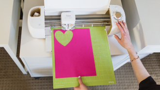 Person's hand holding a Cricut mat cutting out pink hearts on a white Cricut machine.