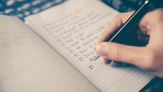 Right hand holding a black pen to a graph paper journal. The journal contains a blurry list with check boxes next to each item. 