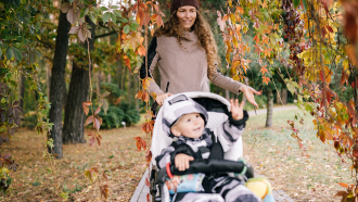 Mom walking with a baby in a stroller on a walking path surrounded by fall leaves. 