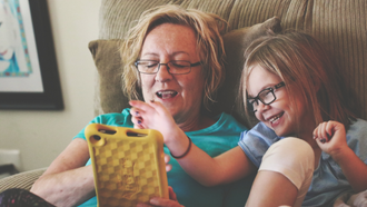 Blonde older woman wearing black framed glasses and a blue shirt sits with a little girl. The little girl is blonde wearing a blue striped shirt and black framed glasses. Both are sitting on a tan corduroy chair leaning on a tan pillow pointing at a yellow tablet. 
