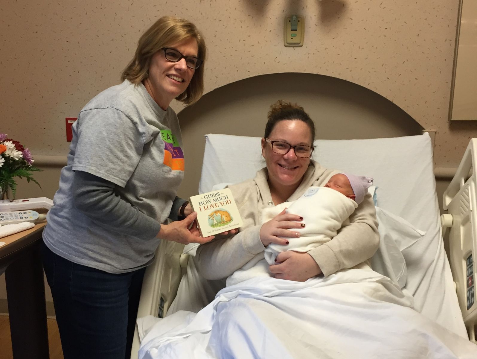 Woman in hospital holding a newborn baby while a library staff member hands her a book for the new baby.